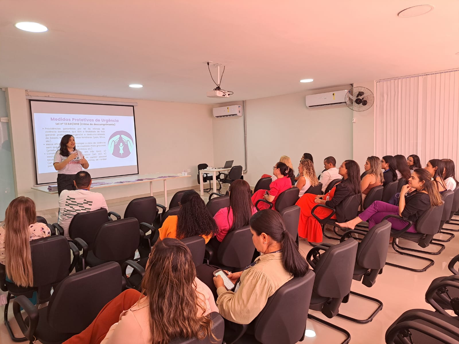 imagem mostra uma sala de aula ou auditório com diversas pessoas sentadas assistindo a uma apresentação. A apresentadora está de pé na frente, ao lado de uma tela de projeção, onde há um slide com o título "Medidas Protetivas de Urgência". O conteúdo do slide explica a Lei 13.641/2019 sobre crime de descumprimento de medidas protetivas, listando algumas providências legais para casos de violência doméstica.

Entre os pontos apresentados no slide, estão:

Afastamento do lar, domicílio ou convivência com a vítima.

Proibição de condutas específicas.

Restrição ou suspensão de visitas aos dependentes menores.

Encaminhamento para programas de proteção ou atendimento.

A sala é bem iluminada, com luzes no teto, ar-condicionado nas paredes, e as pessoas parecem atentas à apresentação, algumas até tomando notas.