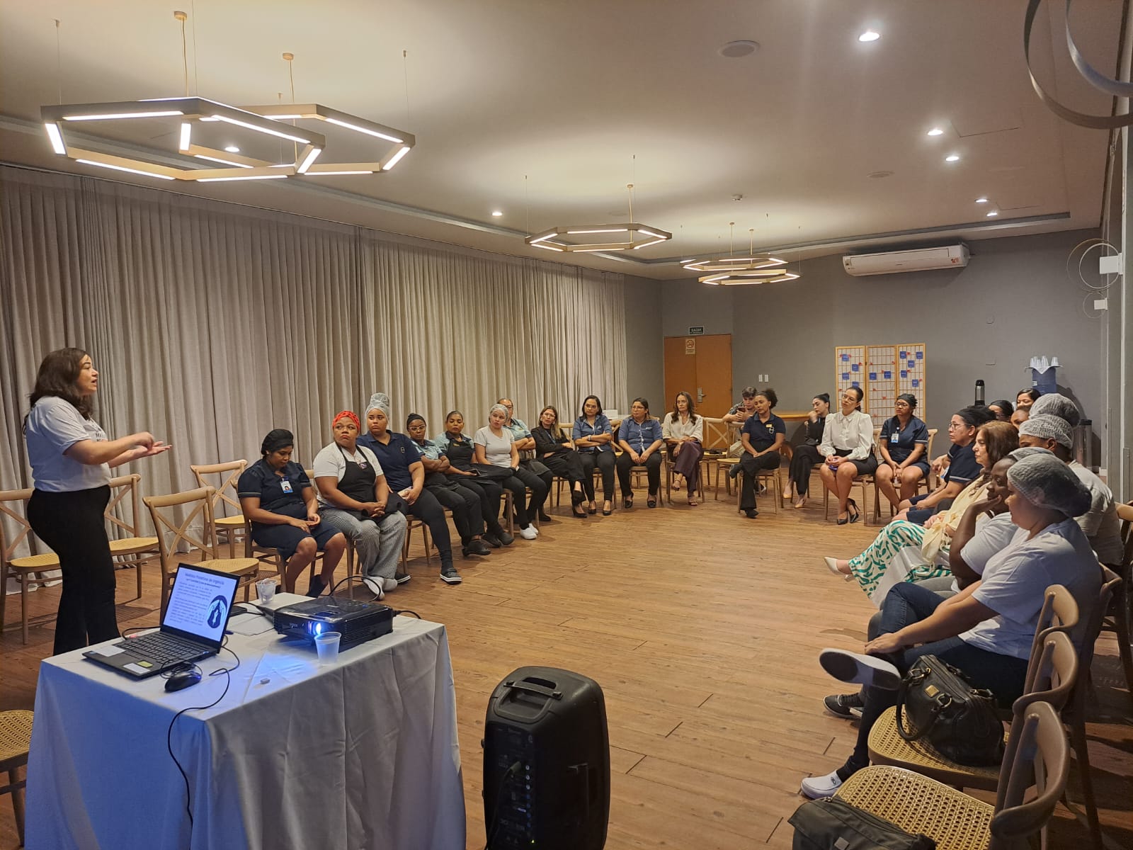 A imagem mostra um grupo de pessoas sentadas em círculo em uma sala, possivelmente para uma reunião, oficina ou sessão de terapia em grupo. Uma mulher está de pé no lado esquerdo da imagem, parecendo se dirigir ao grupo. No primeiro plano, há uma mesa com um laptop, um projetor e alguns papéis. A sala tem piso de madeira, luminárias modernas no teto e uma grande cortina cobrindo a parede ao fundo. Há também uma unidade de ar condicionado na parede e um quadro de avisos com vários papéis fixados nele.