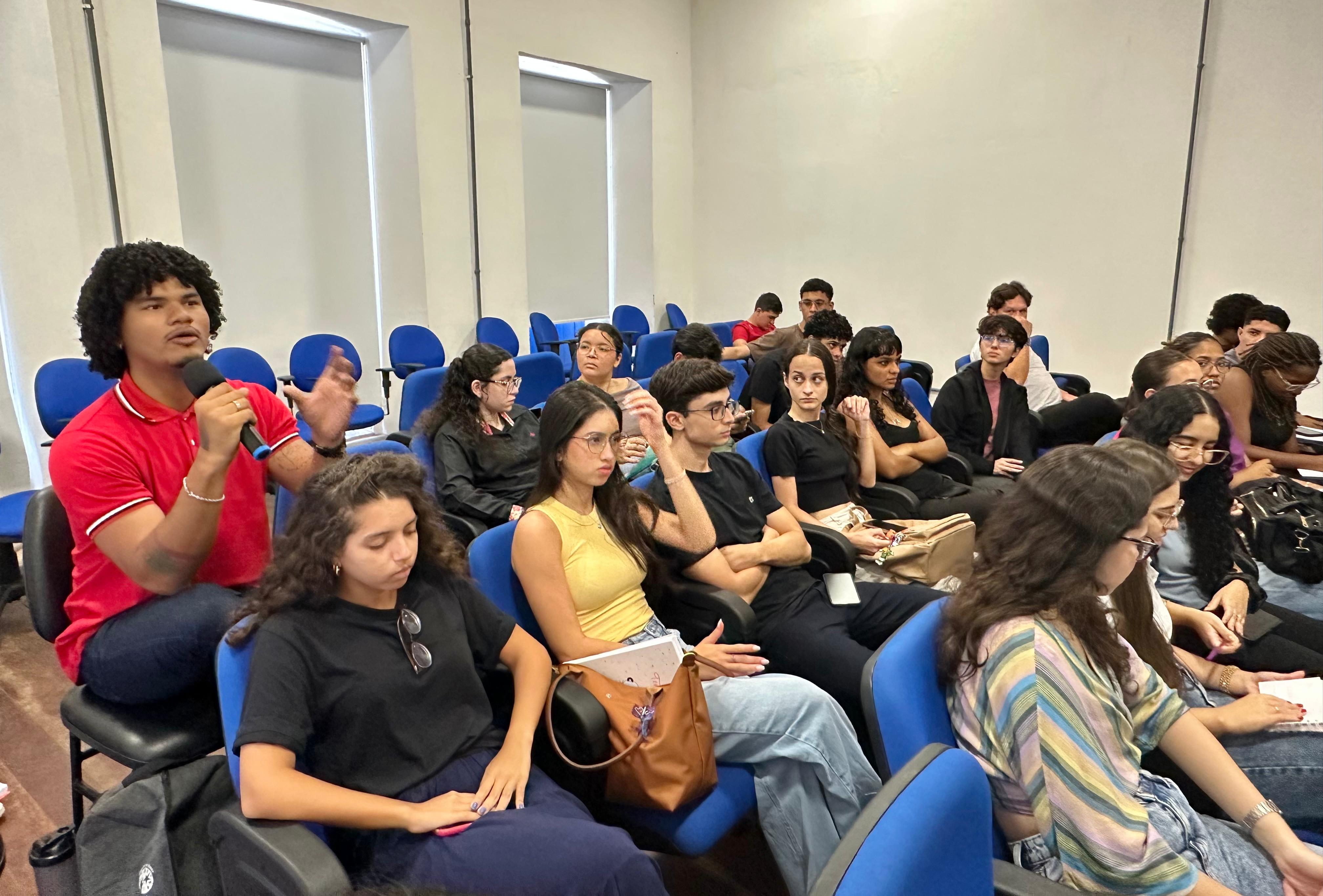 Foto colorida mostra estudantes de Direito da UEMA sentados no auditório. Na imagem um aluno está em pé falando ao microfone.