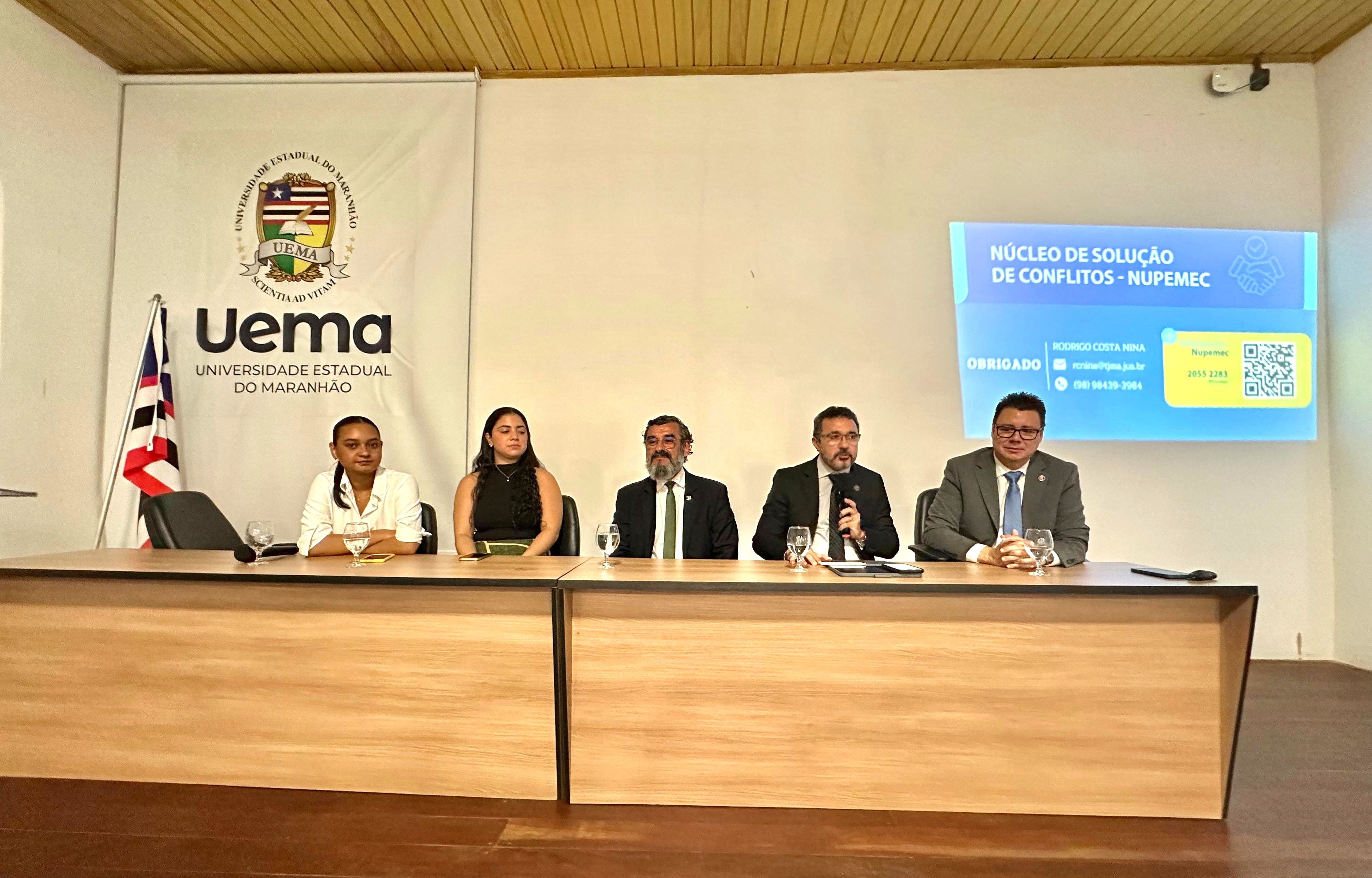 Foto colorida mostra desembargador Nilo sentado em uma mesa falando ao microfone durante palestra na UEMA