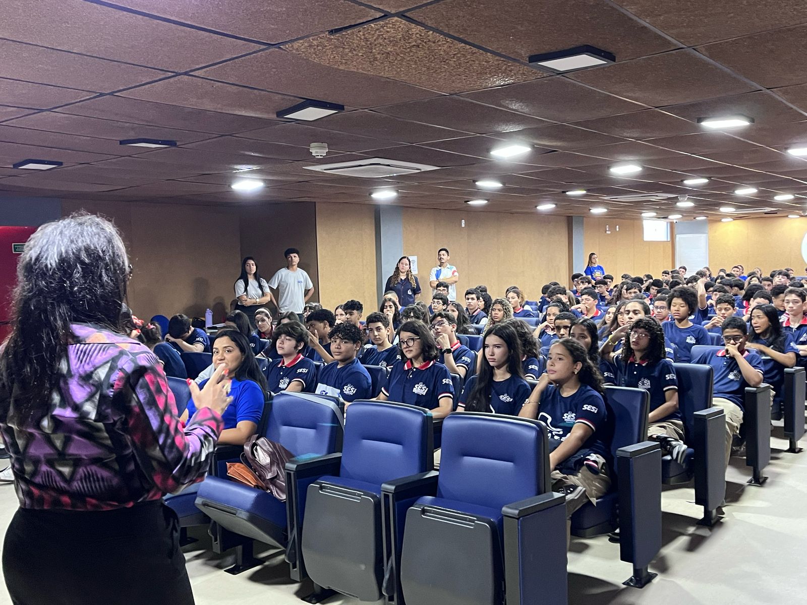 Uma mulher de costas, com cabelos longos e escuros, veste uma blusa estampada roxa, preta e branca. Ela está falando para uma plateia de estudantes que vestem uniformes azul-marinho com a logo "SESI" no peito. Os alunos estão sentados em cadeiras acolchoadas azul-escuras, dispostas em fileiras dentro de um auditório. Alguns estudantes observam atentamente, enquanto outros conversam ou estão distraídos. No fundo, há algumas pessoas de pé, algumas vestindo roupas brancas.