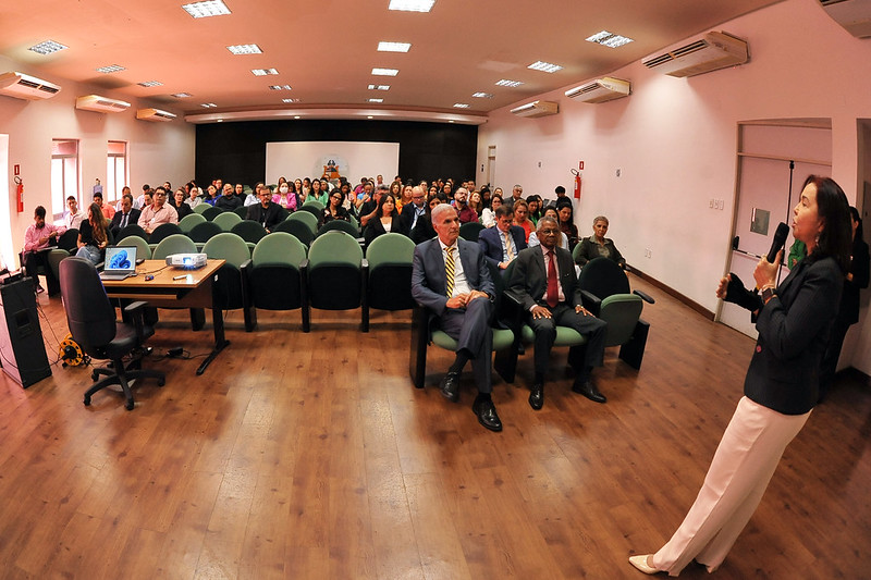 Imagem ampla em ambiente interno de auditorio. Homens e mulheres sentados em cadeiras verdes assistem a fala do professor Gil Pinto, de pé à frente