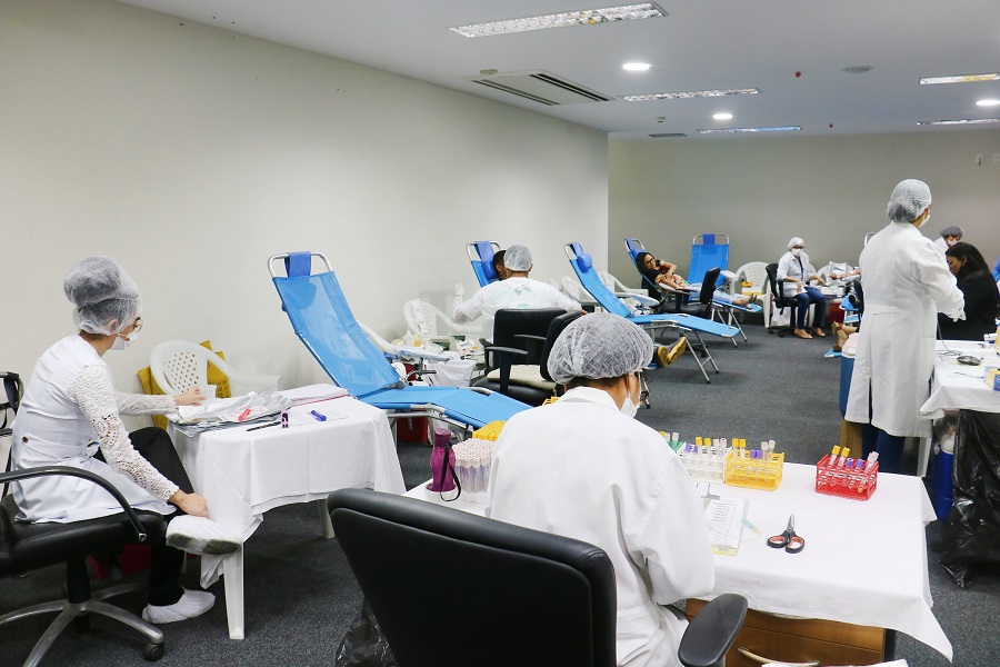 Foto retangular. Auditório com profissionais de saúde em procedimento de doação de sangue. Duas pessoas estão deitadas em cadeiras reclináveis azuis. Elas doam sangue.