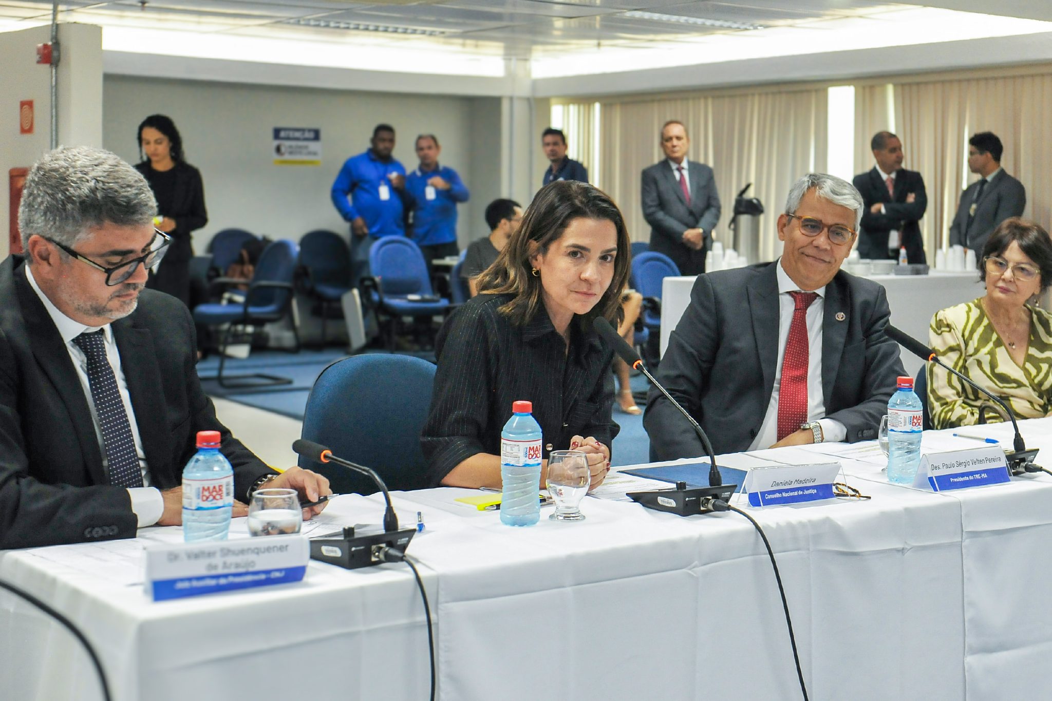 A imagem mostra um ambiente formal onde ocorre uma reunião ou conferência. Há várias pessoas sentadas em torno de uma mesa longa, coberta com uma toalha branca. Na mesa, estão dispostos garrafas de água, copos e microfones, indicando que os participantes estão envolvidos em discussões ou apresentações formais.