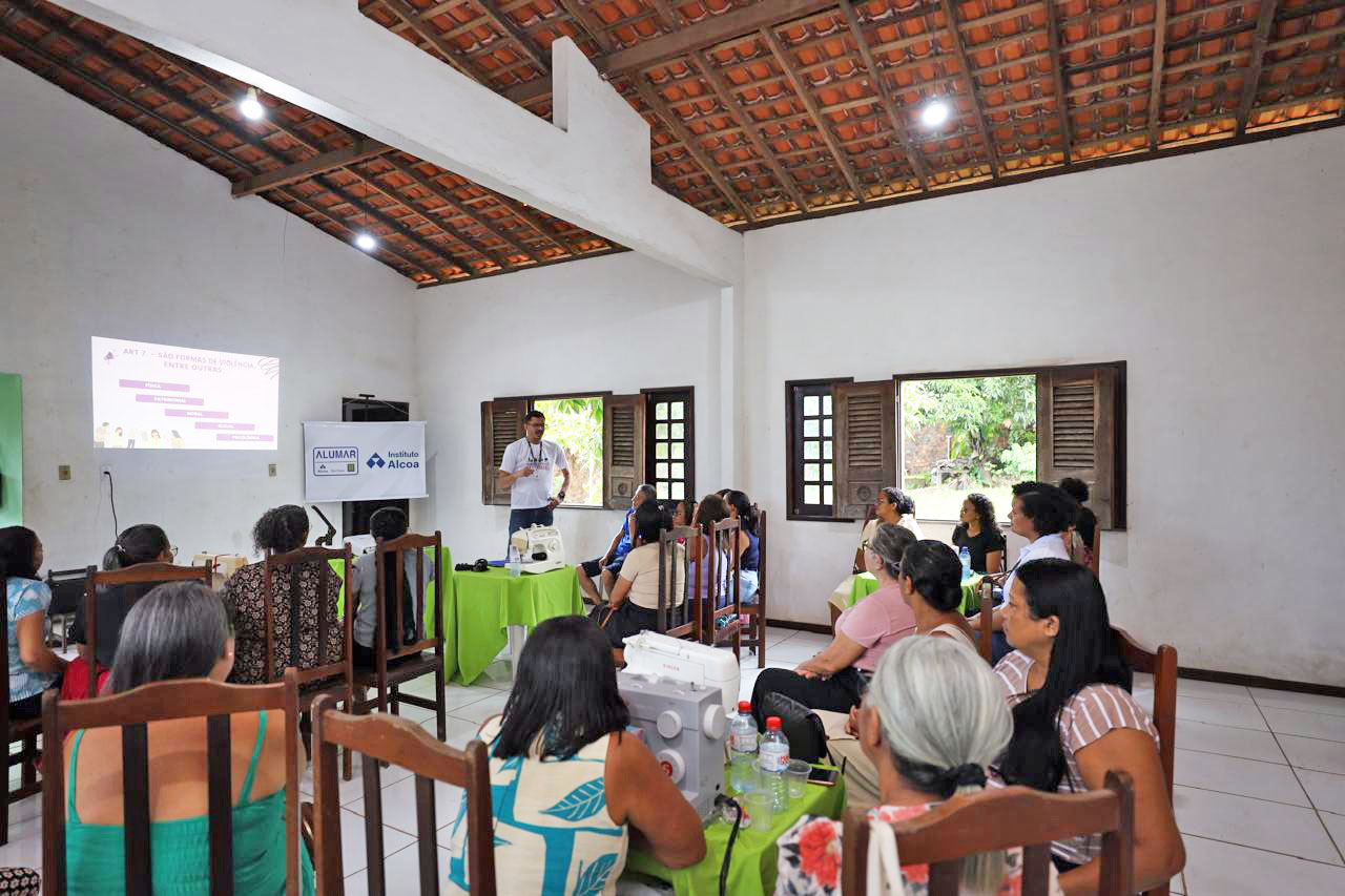 Sala com paredes brancas e telhado de madeira. Um homem apresenta uma palestra sobre formas de violência, com projeção na parede. Mulheres sentadas assistem atentamente. Há máquinas de costura sobre mesas cobertas com toalhas verdes. Banners do Instituto Alcoa e Alumar ao fundo.