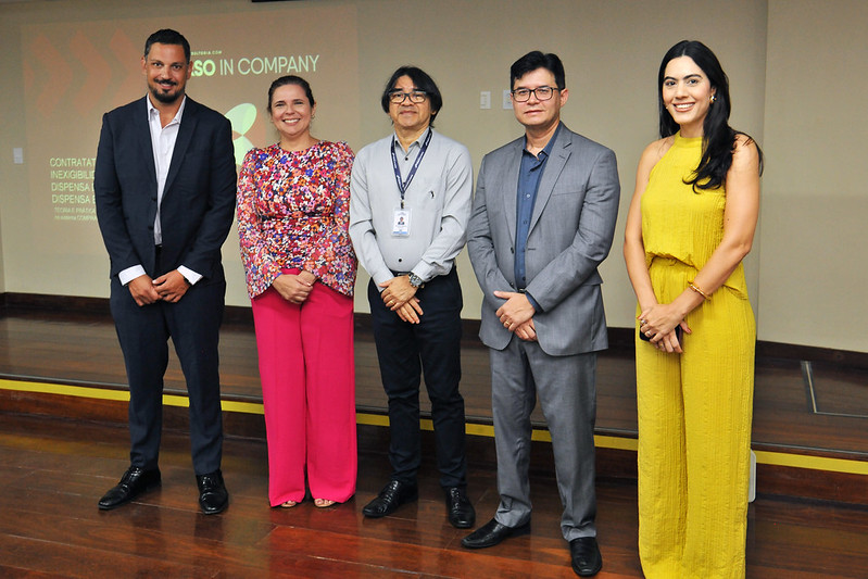 Foto horizontal do Curso de Contratação direta. Nesta foto, cinco pessoas estão em pé, lado a lado, em um palco de madeira. Todas estão vestidas formalmente, com roupas sociais diversas. A partir da esquerda, temos: uma pessoa de terno escuro e sapatos pretos; outra com uma blusa colorida de mangas compridas e calças rosa, trazendo vivacidade ao conjunto; uma terceira pessoa com camisa social clara, calças escuras e um crachá pendurado; logo ao lado, há uma pessoa usando um terno cinza combinado com uma camisa azul; por fim, há uma pessoa vestindo um macacão amarelo e pulseiras chamativas. Ao fundo, há uma projeção na parede com textos parcialmente visíveis, incluindo frases como IN COMPANY e termos como CONTRATAÇÃO, INEXIGIBILIDADE, DISPENSA.