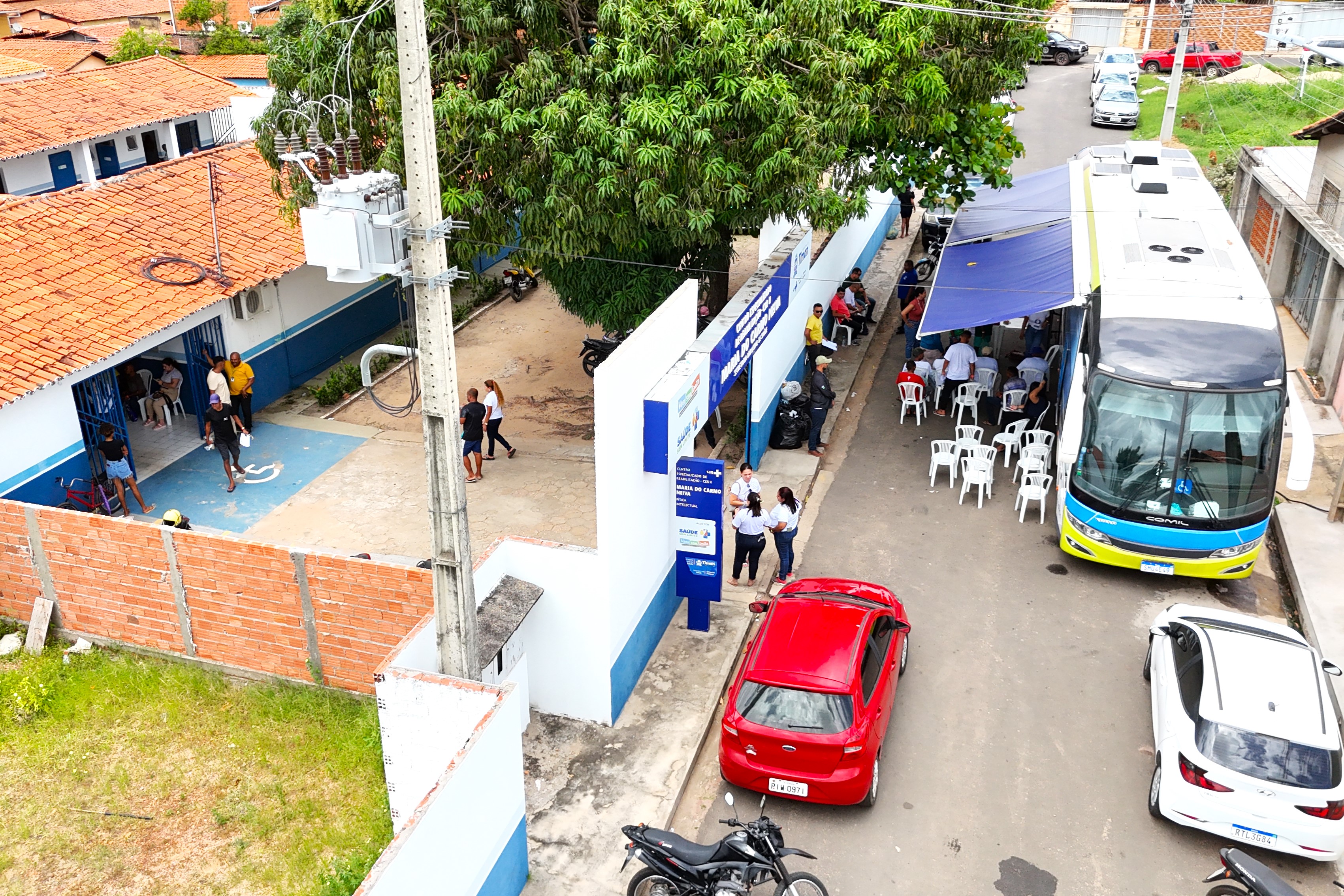 A imagem mostra um grupo de pessoas reunidas em frente a um ônibus identificado com o logotipo "TJMA" (Tribunal de Justiça do Maranhão). O ônibus possui palavras como "Cidadania," "Sociedade," "Processos e Estruturas," "Fiscal" e "Aprendizado e Crescimento" estampadas em sua lateral. Há também um banner que destaca a mensagem "Bota a camisinha, cuide da saúde! Cuide de você." Algumas pessoas estão sentadas em cadeiras plásticas brancas, enquanto outras estão em pé. O cenário sugere um evento ao ar livre, provavelmente ligado à promoção da saúde e cidadania.