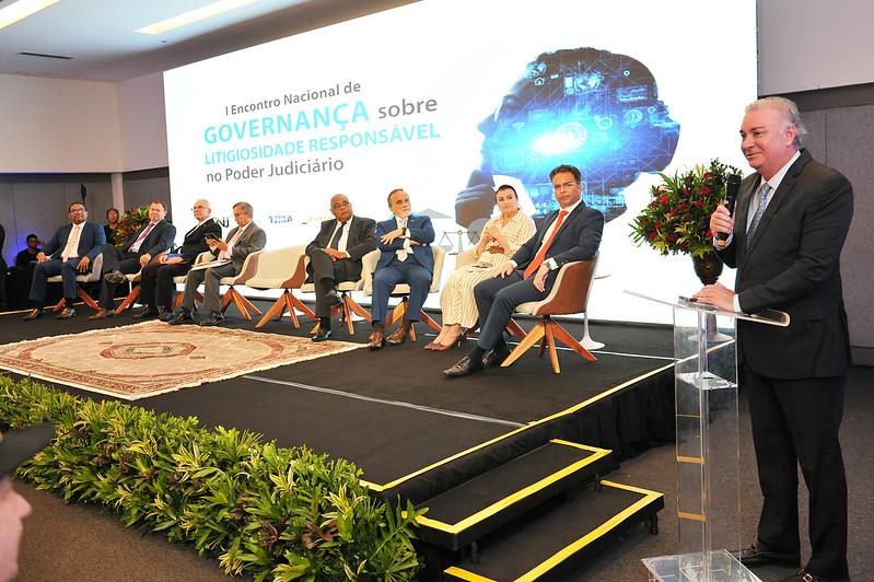 Foto horizontal do I Encontro Nacional sobre Litigiosidade Responsável - presidente. A imagem retrata um evento formal no palco. Há várias pessoas sentadas em cadeiras no fundo, vestidas formalmente com ternos e vestidos. À frente, o presidente do TJMA, desembargador Froz Sobrinho, está falando em um púlpito, sendo o centro da ação. No fundo do palco, um grande painel apresenta o texto: "I Encontro Nacional de GOVERNANÇA sobre LITIGIOSIDADE RESPONSÁVEL no Poder Judiciário". O cenário inclui um tapete central e plantas decorativas ao redor do palco.