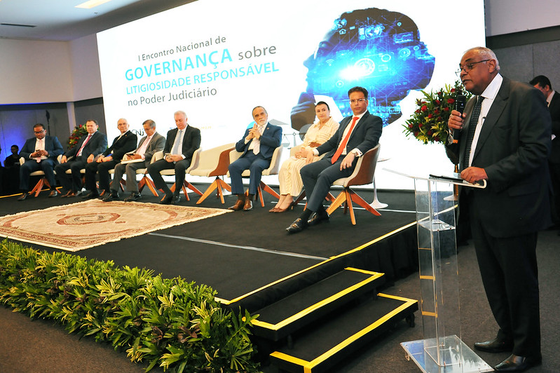Foto horizontal do I Encontro Nacional sobre Litigiosidade Responsável -presidente do Colégio Permanente de Corregedores. A imagem apresenta um evento formal acontecendo em um palco. No lado direito, o desembargador Gilberto Barbosa está de pé falando em um púlpito transparente, provavelmente fazendo um discurso ou apresentação. Atrás dele, há várias pessoas sentadas em cadeiras no palco, todas vestidas formalmente, o que reforça o tom profissional da ocasião. No fundo, um grande painel exibe o texto: "I Encontro Nacional de Governança sobre Litigiosidade Responsável no Poder Judiciário". O painel também possui uma imagem gráfica destacando um perfil humano com elementos tecnológicos ao redor. Há plantas decorativas na frente do palco, contribuindo para a composição elegante do ambiente. Tudo aponta para uma conferência ou reunião institucional de grande importância.