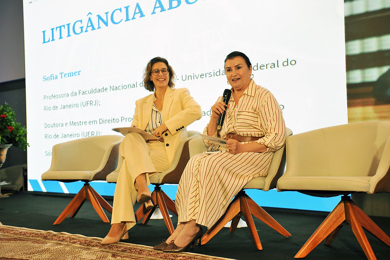 Foto horizontal do I Encontro Nacional  sobre Litigiosidade Responsável - presidente do Grupo Maria Firmina. A imagem apresenta duas pessoas sentadas em um palco, participando de um evento formal. Uma delas está segurando um microfone e parece estar falando. Atrás delas, há um grande painel com o seguinte texto em destaque: "LITIGÂNCIA ABOLITIVA" "Sofia Temer": Professora da Faculdade Nacional de Direito - Universidade Federal do Rio de Janeiro (UFRJ); Doutora e Mestre em Direito Processual pela Universidade Federal do Rio de Janeiro (UFRJ). O palco é decorado com cadeiras modernas e um design profissional.