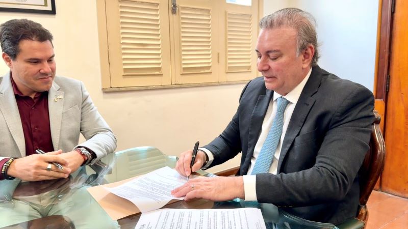 Foto horizontal de assinatura de parceria TáxiGov entre TJMA e Mapa.Na foto, o presidente do TJMA e o presidente da Mapa assinam documento, sentados em frente a uma mesa com tampo de vidro. O presidente veste um terno preto com uma gravata azul clara. Ele está concentrado na ação de assinar o documento com uma caneta. À esquerda, o presidente da Mapa usa um blazer cinza claro e uma camisa vermelha, segurando uma caneta, aguardando sua vez de assinar. Sobre a mesa, há um envelope pardo, parcialmente coberto pelo documento que está sendo assinado. O espaço ao fundo é iluminado e apresenta uma janela com persianas fechadas, complementada por uma parede clara.