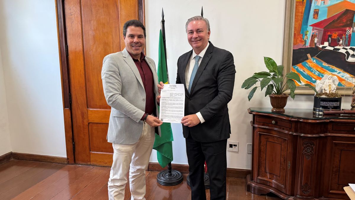 Foto horizontal de encontro sobre uso TáxiGov pelo TJMA. foto mostra o presidente do TJMA e o presidente da Mapa, em pé. Eles estão segurando um documento oficial. Atrás, há uma bandeira verde e, ao lado, um móvel com uma planta e um objeto decorativo. Na parede, há uma pintura colorida com elementos arquitetônicos e figuras. Um ambiente bem formal e representativo, no gabinete da Presidência do TJMA.