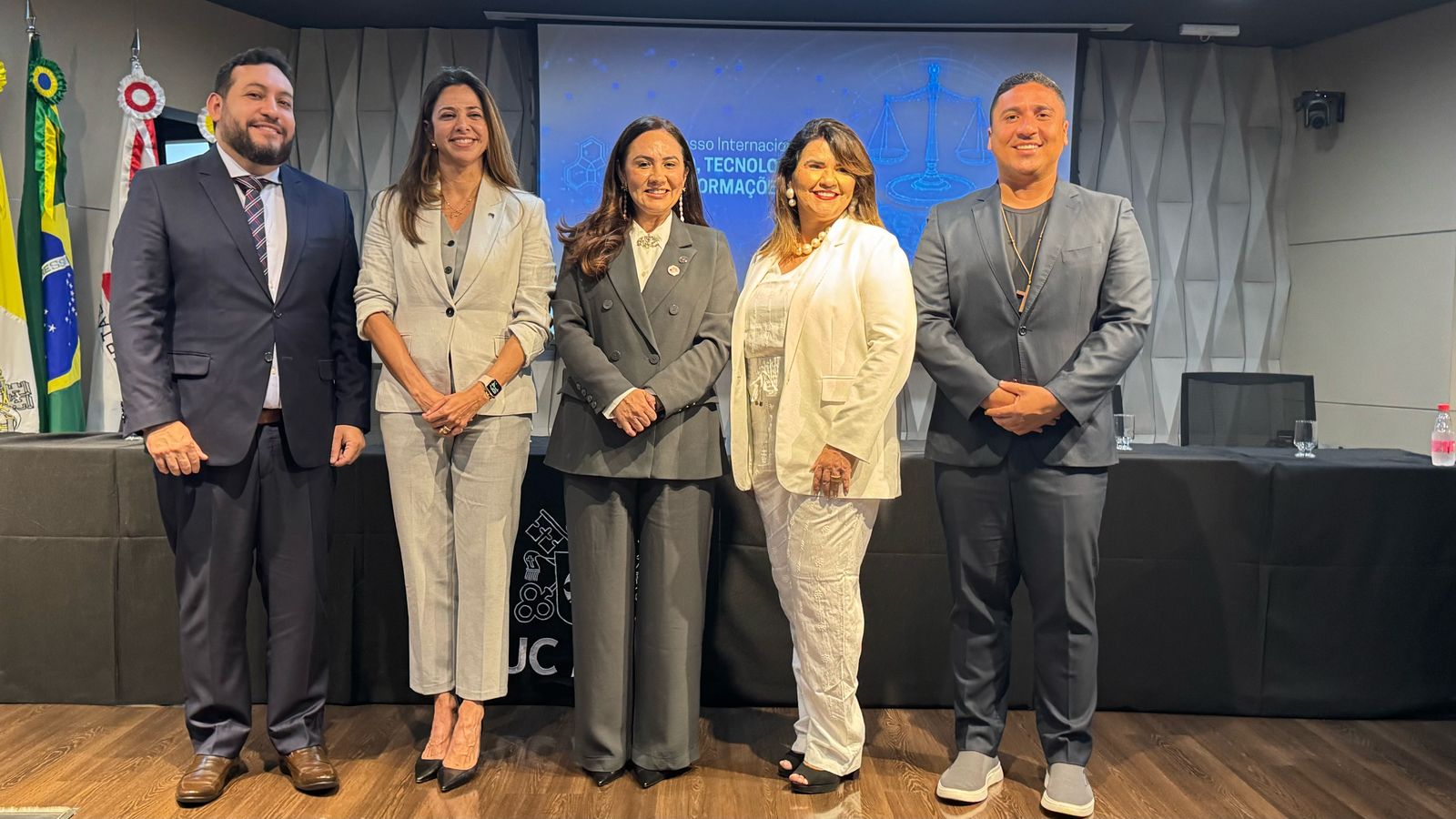 A imagem apresenta cinco pessoas em pé, lado a lado, em frente a uma mesa coberta com uma toalha preta. O ambiente é formal, sugerindo um evento acadêmico ou institucional. As pessoas vestem roupas sociais, como ternos e vestidos, transmitindo um tom de seriedade e profissionalismo.

Ao fundo, há uma tela de projeção exibindo o texto "Congresso Internacional de Tecnologia da Informação", acompanhado da imagem de uma balança de justiça, reforçando uma possível conexão entre direito e tecnologia. À esquerda da tela, encontram-se quatro bandeiras, incluindo a do Brasil, que ajudam a compor o cenário oficial do evento.

Sobre a mesa, há alguns copos de água e uma garrafa vermelha, indicando que os participantes podem estar prestes a falar ou já participaram de um momento de discussão. O chão é de madeira e as paredes apresentam um design geométrico, adicionando um toque moderno ao espaço.