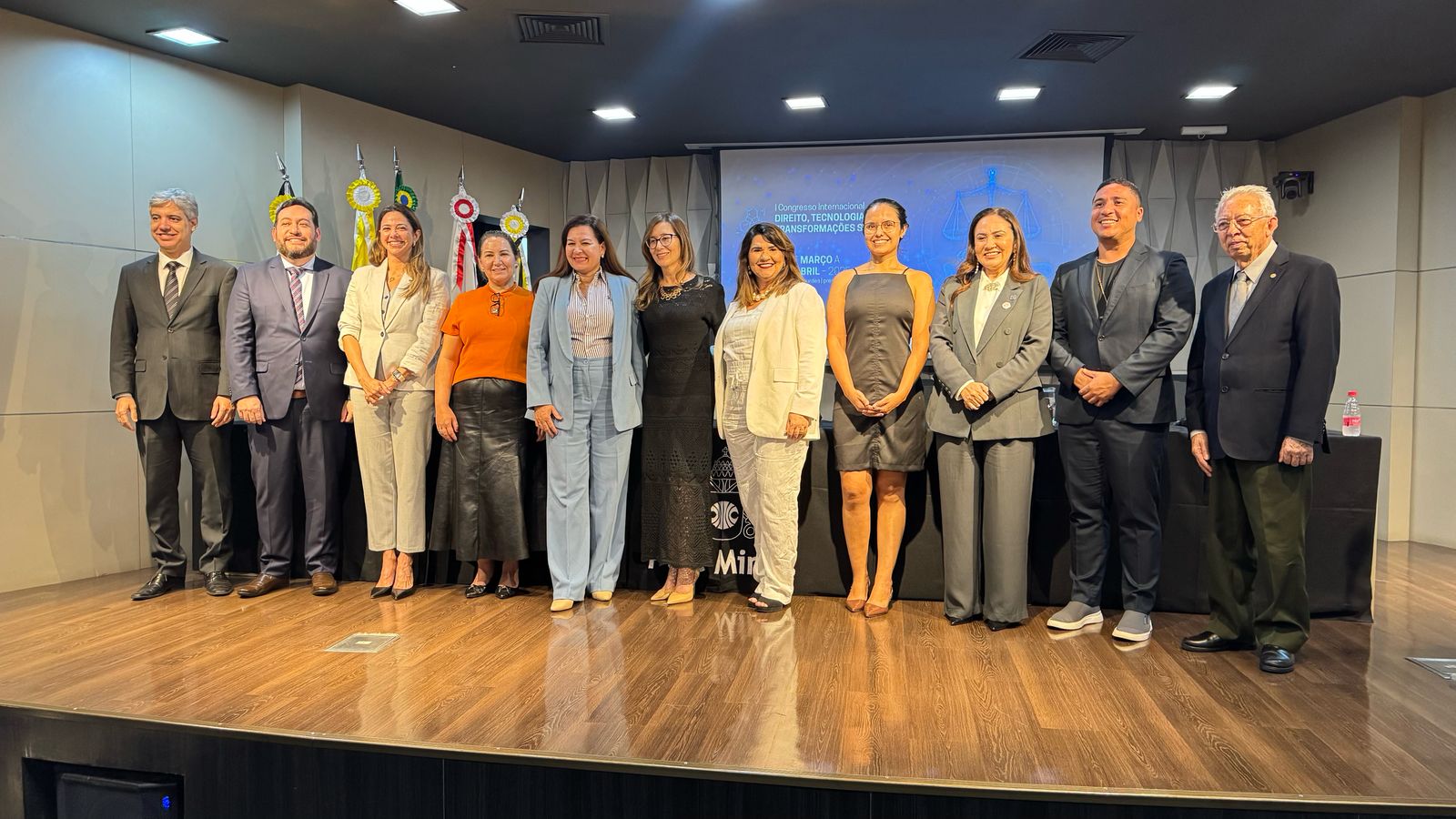 A imagem mostra um grupo de doze pessoas em pé em um palco, lado a lado, posando para uma foto. O palco é de madeira e o fundo é composto por uma parede clara com algumas bandeiras e um painel de projeção. As pessoas estão vestidas formalmente, com ternos, vestidos e roupas sociais. No fundo, há um painel de projeção que exibe o texto "I Congresso Internacional de Direito, Tecnologia e Transformações Sociais" e as datas "30 de março a 1 de abril".