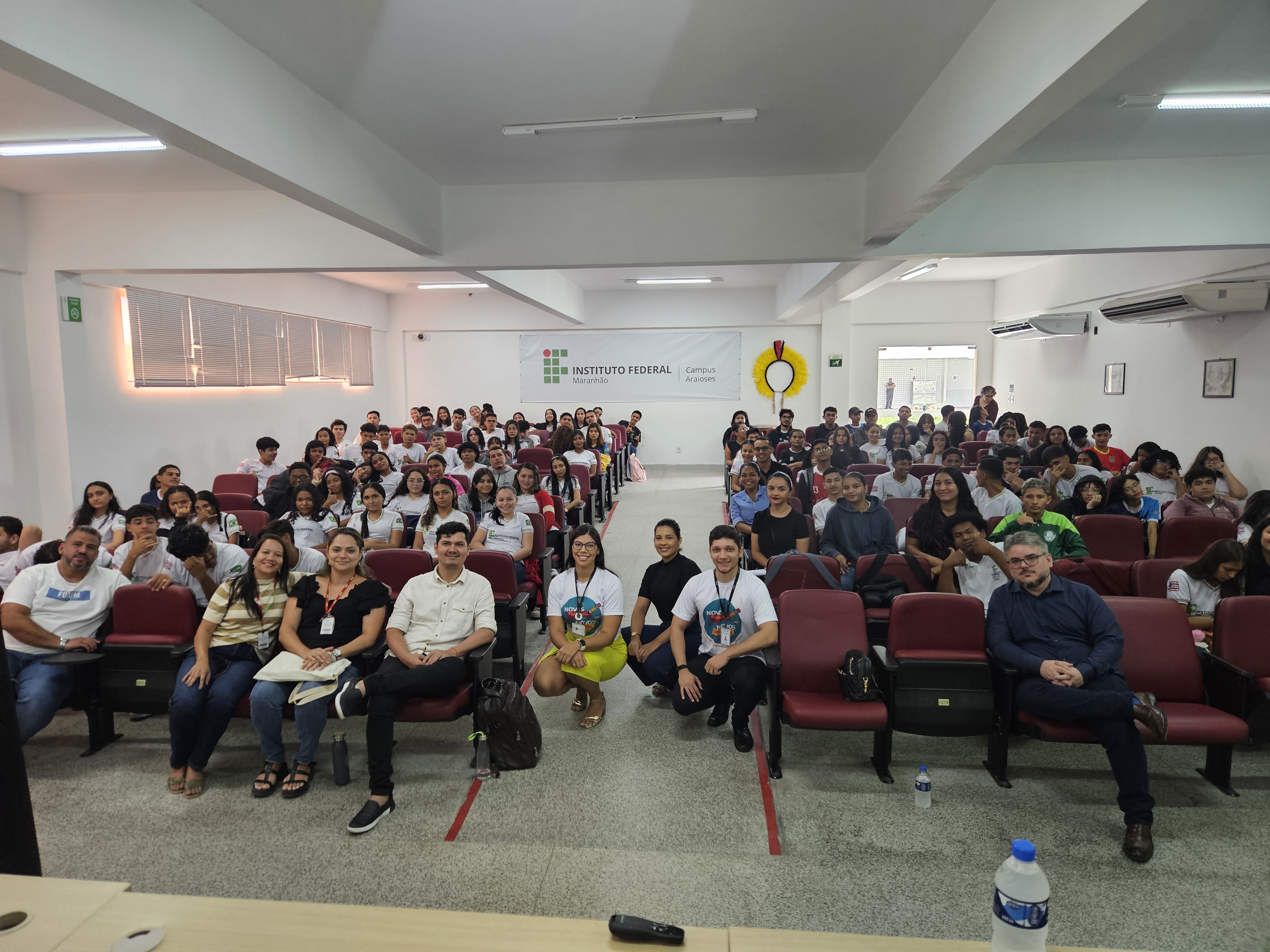 Um auditório grande, do Instituto Federal do Maranhão, Campus Araioses, repleto de alunos uniformizados e professores. Eles estão sentados em cadeiras vermelhas, organizados em fileiras. Na frente, um grupo de pessoas está em pé e sentada, sorrindo para a foto. A parede ao fundo tem a identificação do Instituto e decoração com um adereço indígena em amarelo e vermelho. O ambiente é claro, com iluminação artificial e janelas laterais com persianas.