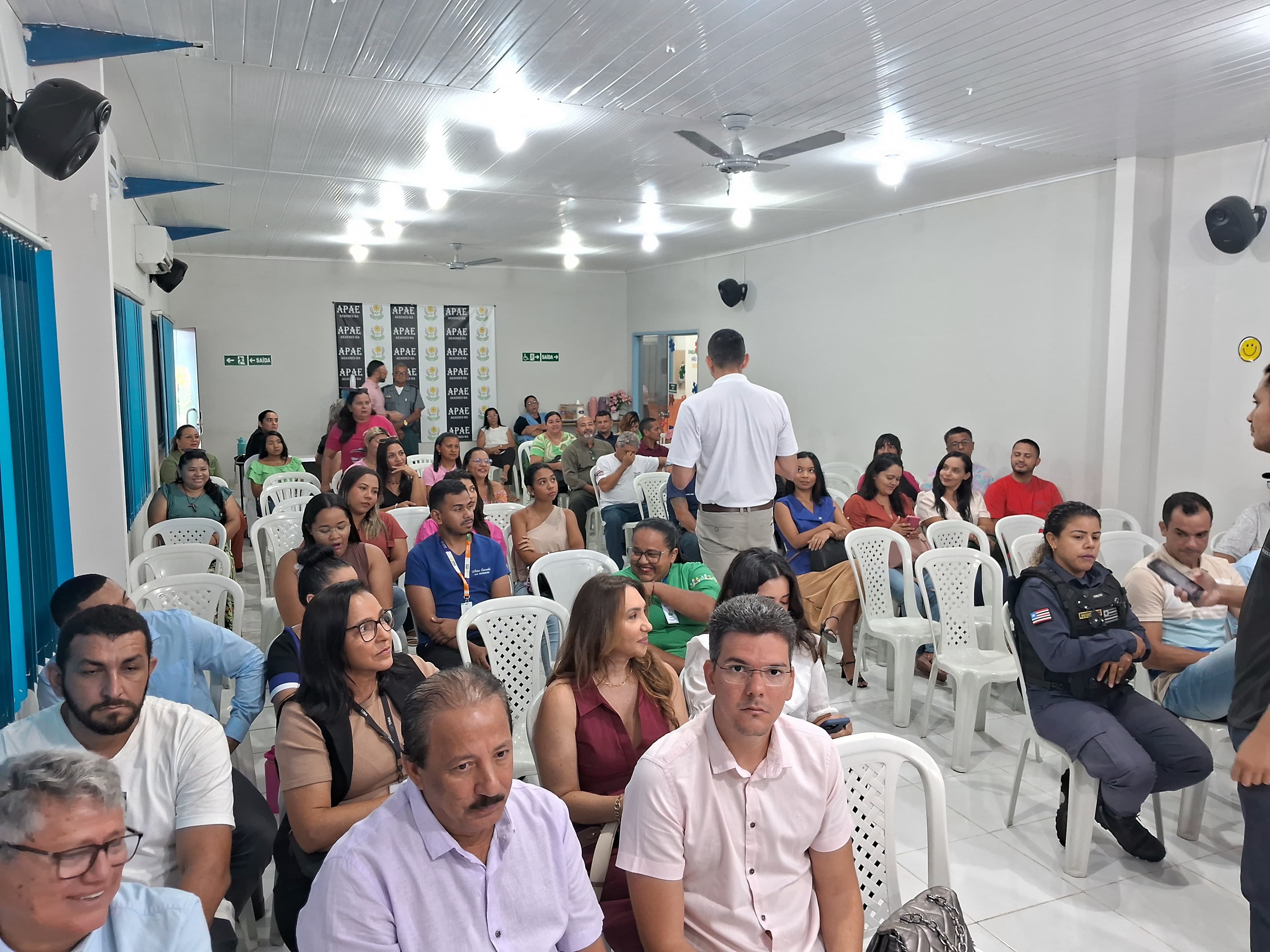 Um auditório amplo, com diversas cadeiras brancas de plástico, está cheio de pessoas sentadas, distribuídas de maneira organizada. Há uma mistura de homens e mulheres, alguns fardados (polícia), outros com crachás, e muitas pessoas com expressões atentas. Na parte de trás do auditório, há banners da APAE fixados nas paredes. O ambiente é claro, com teto de forro branco, iluminação de lâmpadas brancas, ventiladores de teto, e paredes claras.