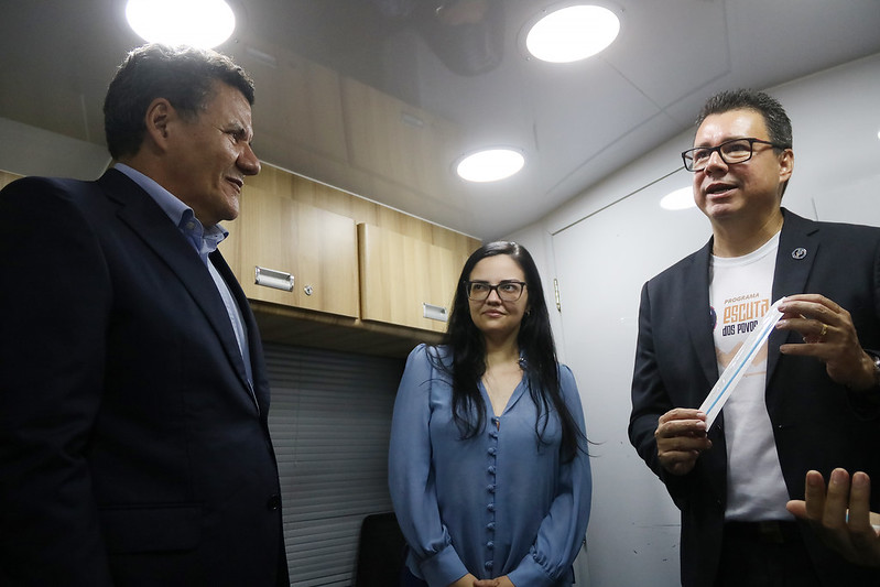 Foto horizontal. Novo recorde no mutirão de cidadania indígena 3.476 atendimentos em Grajaú. A foto mostra três pessoas interagindo em um espaço interno pequeno dentro de um ônibus. À esquerda, um homem de pele clara veste um terno azul escuro sobre uma camisa azul clara. Ele está olhando para a direita e sorrindo levemente. No centro, uma mulher de pele clara com cabelos escuros longos e óculos está olhando para o homem à sua esquerda e sorrindo. Ela veste uma blusa azul de mangas compridas. À direita, um homem de pele clara veste um blazer preto sobre uma camiseta branca com texto e um logotipo. Ele usa óculos e está olhando para a direita, gesticulando com as mãos enquanto fala.