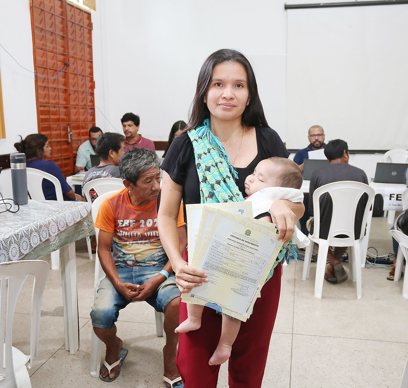 Foto horizontal. Novo recorde no mutirão de cidadania indígena 3.476 atendimentos em Grajaú. A foto mostra uma mulher de pele morena clara em primeiro plano, segurando um bebê que parece estar dormindo e vários documentos de certidões. A mulher veste uma blusa preta, calças vinho e tem um tecido estampado em tons de azul e amarelo usado como um tipo de para segurar o bebê. Ela olha para a câmera com uma expressão calma. Ao fundo, há várias pessoas sentadas em mesas brancas, algumas com laptops.  O ambiente é uma sala com paredes claras e uma porta de madeira à esquerda. Uma tela de projeção branca está pendurada na parede ao fundo.