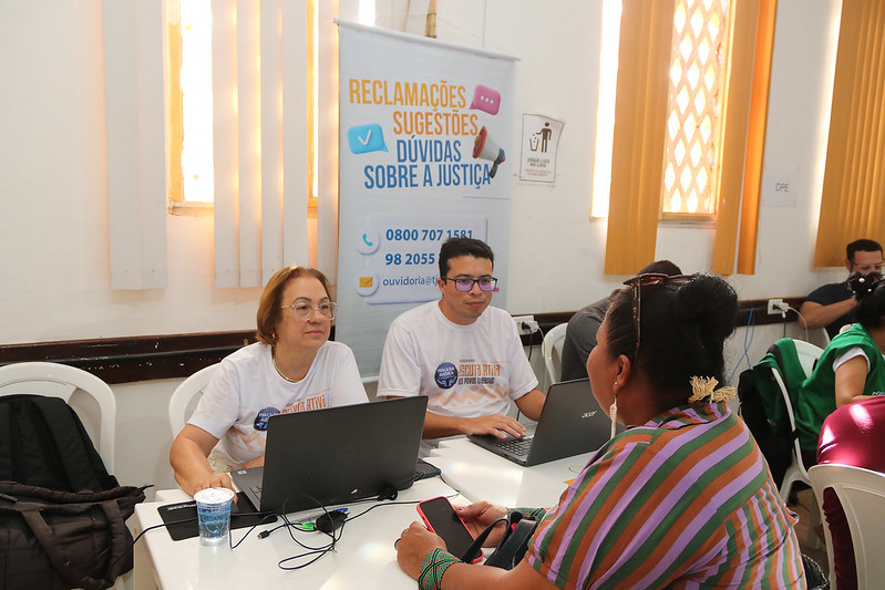 Foto horizontal. Novo recorde no mutirão de cidadania indígena 3.476 atendimentos em Grajaú. A foto mostra três pessoas interagindo em um ambiente de uma sala de informações. À esquerda, uma mulher mais velha com óculos e cabelos curtos veste uma camiseta branca com uma estampa e está olhando para a tela de um laptop preto sobre uma mesa branca. Ao centro, um homem jovem com óculos veste uma camiseta branca com a mesma estampa e também está olhando para a tela de um laptop preto sobre a mesma mesa. Há um banner atrás dele com texto em português, incluindo 