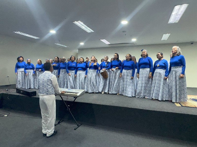 Foto horizontal do PPA do TJMA. A foto mostra um coral, composto principalmente por mulheres, em um palco. Elas estão vestindo uniformes que consistem em blusas azuis de manga comprida e saias longas com um padrão azul e branco. Há um homem de costas para a câmera, também vestido com um padrão azul e branco, que está conduzindo o coral e tocando um teclado musical à sua frente. As pessoas no coral parecem ser de idade avançada.