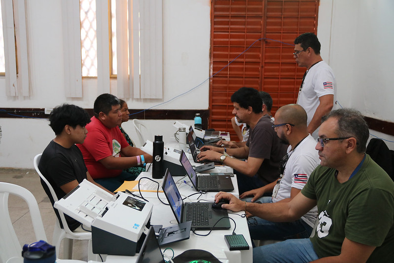 Foto horizontal de atendimento em que o Programa Praça da Justiça oferece serviços gratuitos a povos indígenas de Grajaú. A foto mostra um grupo de sete pessoas em um ambiente de uma sala com várias mesas brancas dispostas em fileira. A sala tem paredes claras e janelas com grades. As pessoas estão sentadas em cadeiras brancas de plástico, trabalhando em laptops que estão sobre as mesas. Há cinco laptops visíveis, e algumas pessoas parecem estar digitando ou olhando para as telas. Há também alguns outros objetos sobre as mesas, como garrafas de água, papéis e uma impressora.  As pessoas na foto parecem ser adultos de diferentes idades. Suas roupas são casuais, incluindo camisetas e camisas. Suas expressões faciais variam, com alguns concentrados em seus trabalhos e outros interagindo ou observando.