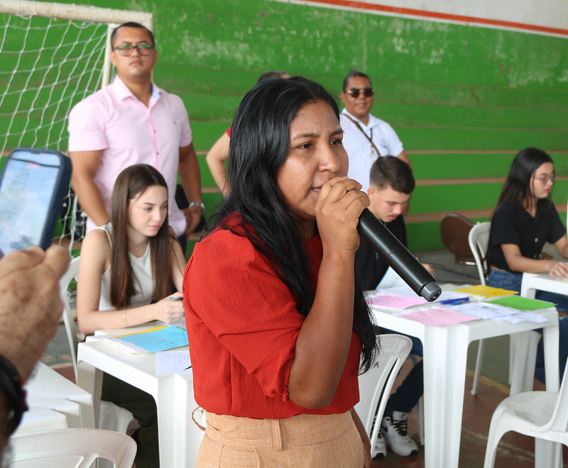 Foto horizontal de atendimento em que o Programa Praça da Justiça oferece serviços gratuitos a povos indígenas de Grajaú. A foto mostra uma mulher de pele morena e cabelos longos e pretos falando em um microfone. Ela veste uma blusa vermelha de mangas curtas e uma saia ou calça de cor bege clara. Ela está fazendo um anúncio. Ao fundo, há várias pessoas sentadas em mesas brancas. À esquerda, uma jovem de pele clara com cabelos longos e castanhos está sentada e olhando para baixo. Atrás dela, um homem de pele clara com óculos e uma camisa rosa está em pé, olhando para a frente. À direita da mulher que fala, há um jovem de pele clara com cabelos escuros e uma camiseta preta. Mais à direita, uma jovem de pele clara com cabelos escuros e óculos está sentada a uma mesa, olhando para algo em sua frente. Atrás dela, um homem de pele morena clara com óculos de sol e uma camisa branca está em pé. O ambiente é um ginásio.