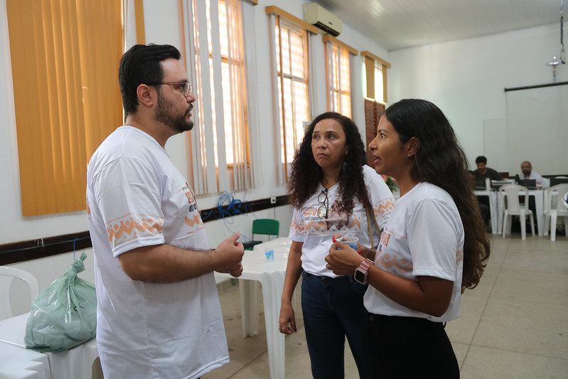 Foto horizontal de atendimento em que o Programa Praça da Justiça oferece serviços gratuitos a povos indígenas de Grajaú. A foto mostra três pessoas em primeiro plano, envolvidas em uma conversa em um ambiente interno que parece ser uma sala de eventos ou um espaço comunitário. À esquerda, um homem de pele clara com barba escura e óculos veste uma camiseta branca com uma estampa em tons de laranja e azul. Ele está de perfil, olhando e gesticulando enquanto fala com as duas mulheres à sua direita. No centro, uma mulher de pele morena clara com cabelos cacheados escuros veste uma camiseta branca com a mesma estampa do homem e calças jeans azuis. Ela está olhando para o homem e parece estar ouvindo atentamente. À direita, uma mulher de pele morena clara com longos cabelos escuros veste a mesma camiseta branca e calças pretas. Ela está olhando para o homem e parece estar falando ou respondendo. Ela segura um copo descartável na mão direita. Ao fundo, a sala tem paredes claras e grandes janelas com cortinas finas que deixam passar a luz natural.