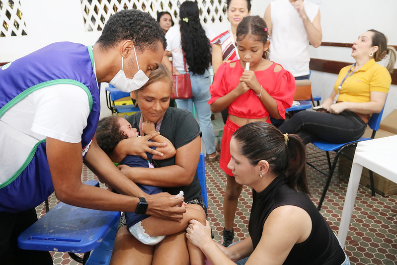 Foto horizontal em que o Programa Praça da Justiça oferece serviços gratuitos a povos indígenas de Grajaú. A foto mostra uma cena em um ambiente de posto de saúde e local de vacinação temporário. No centro da imagem, um profissional de saúde do sexo masculino, vestindo uma máscara facial branca, uma camiseta branca e um colete azul com detalhes verdes, está aplicando uma injeção na perna de um bebê pequeno que está sendo segurado por uma mulher. A mulher, que aparenta ser a mãe do bebê, está sentada em uma cadeira azul e parece estar tentando acalmar a criança que está chorando. À direita, outra profissional de saúde do sexo feminino, vestindo uma blusa preta de gola alta e com o cabelo preso, parece estar preparando ou auxiliando no procedimento. Ela está olhando para a perna do bebê. Em volta, há outras pessoas esperando ou observando. Uma menina com uma blusa vermelha e uma máscara no queixo está em pé perto da mulher que segura o bebê. Ao fundo, há outras mulheres e uma pessoa vestindo uma camiseta branca. Uma mulher vestindo uma blusa amarela está sentada em uma cadeira azul à direita, olhando para a cena.