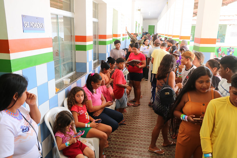 Foto horizontal do Projeto de Cidadania Indígena do TJMA em Grajaú. A imagem mostra um corredor de uma escola, onde muitas pessoas, incluindo crianças e adultos, estão esperando ou conversando. As paredes do corredor são decoradas com azulejos em tons de azul claro e branco, com faixas horizontais verdes e laranjas. Na parede, há uma placa que indica "Gestão Pedagógica". Algumas pessoas estão sentadas em cadeiras de plástico branco, enquanto outras permanecem em pé.