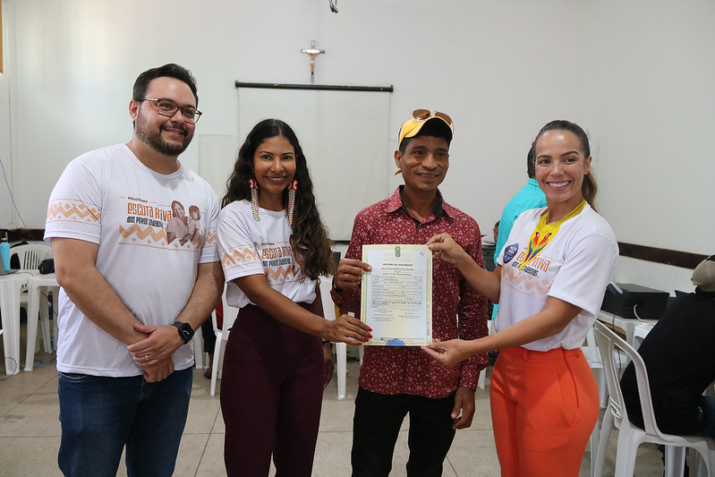 Foto horizontal do Projeto de Cidadania Indígena do TJMA em Grajaú. A foto mostra quatro pessoas em um ambiente interno, posando para uma foto enquanto duas delas entregam um certificado para outra. Da esquerda para a direita: Um homem de pele clara com barba escura e óculos veste uma camiseta branca com uma estampa em tons de marrom e azul, e calças jeans azuis. Ele está sorrindo para a câmera com os braços cruzados. Uma mulher de pele morena clara com longos cabelos escuros e brincos longos e coloridos veste uma camiseta branca com a mesma estampa do homem à esquerda e calças vinho. Ela está sorrindo e olhando para a pessoa que recebe o certificado. Um homem de pele morena veste uma camisa de manga comprida estampada em tons de vermelho e preto, um boné amarelo e óculos de sol na cabeça. Ele está sorrindo e segurando o certificado com as duas mãos. Uma mulher de pele clara com cabelos castanhos presos veste uma camiseta branca com a mesma estampa e calças laranja. Ela está sorrindo e entregando o certificado junto com a mulher à sua esquerda. Ela também usa um cordão com um crachá no pescoço.