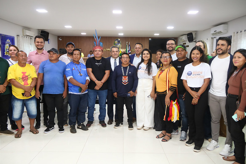 Foto horizontal da Solenidade de abertura do Programa Praça da Justiça em Grajaú. A imagem mostra um grupo de 17 pessoas reunidas em um ambiente fechado, no Plenário da Câmara Municipal de Grajaú. Todas estão de pé, organizadas em duas fileiras. As vestimentas variam entre roupas casuais e formais. Ao fundo, há uma parede decorada com bandeiras e um brasão. Também há uma mesa, atrás da qual algumas pessoas estão sentadas. A iluminação é clara, com várias luzes no teto garantindo visibilidade.