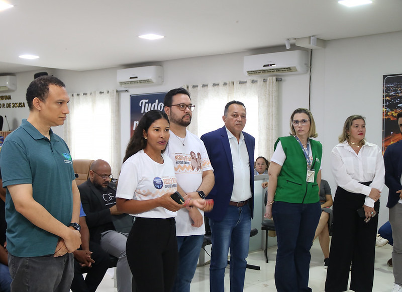 Foto horizontal da Solenidade de abertura do Programa Praça da Justiça em Grajaú. A imagem mostra um grupo de seis pessoas em primeiro plano, em um ambiente interno que parece ser uma sala de reuniões ou um espaço de eventos. Da esquerda para a direita: Um homem de pele morena clara e cabelos curtos e escuros veste uma camisa polo verde azulada e calças cáqui. Ele olha para a esquerda com uma expressão séria.
Uma mulher de pele morena clara e cabelos longos e castanhos veste uma camiseta branca com uma estampa colorida e calças pretas, com a boca aberta e as mãos gesticulando. Um homem de pele clara, com barba e bigode escuros e óculos, veste uma camiseta branca com uma estampa e uma faixa azul e branca sobre o ombro. Um homem de pele clara e cabelos curtos e escuros veste um blazer azul sobre uma camisa branca com listras finas e calças jeans azuis. Uma mulher de pele clara e cabelos loiros curtos veste uma camiseta cinza, um colete verde com um cordão no pescoço e calças jeans azuis. Uma mulher de pele clara e cabelos loiros na altura dos ombros veste uma blusa branca e uma saia preta. Ao fundo, parcialmente visíveis, há outras pessoas sentadas.