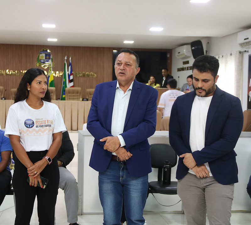 Foto horizontal da Solenidade de abertura do Programa Praça da Justiça em Grajaú. A imagem mostra três pessoas em primeiro plano, em um auditório. À esquerda, uma mulher de pele morena clara e cabelos escuros veste uma camiseta branca com uma estampa em tons de laranja e preto, calças pretas e um relógio rosa no pulso esquerdo. Ela olha para a direita com uma expressão séria e segura um celular na mão direita. No centro, um homem de pele clara, com cabelos escuros e curtos, veste um blazer azul sobre uma camisa branca e calças jeans azuis. Ele está com as mãos juntas à frente do corpo, com a boca ligeiramente aberta e os olhos fixos em algo fora do campo de visão à sua frente. À direita, outro homem de pele clara, com barba escura e cabelos escuros penteados para trás, veste um blazer azul sobre uma camisa branca e calças cinzas. Ele está com a cabeça ligeiramente baixa e as mãos juntas à frente do corpo, com uma expressão concentrada.  Ao fundo, há várias cadeiras de madeira escuras dispostas em fileiras, algumas ocupadas por outras pessoas.
