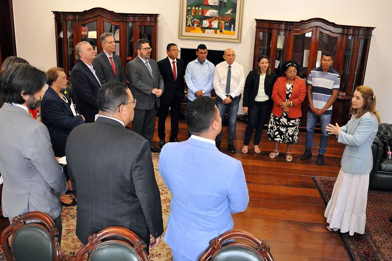 Foto horizontal em que o TJMA celebra acordo para solução de conflitos na Comunidade Jussaral. A foto mostra um grupo de pessoas em uma sala, no Gabinete da Presidência do TJMA . Há vários homens e mulheres, a maioria em pé, alguns observando a deputada à direita que está falando, gesticulando com as mãos. As pessoas estão vestindo roupas sociais. Ao fundo, há móveis de madeira escura, como estantes ou armários, e um quadro colorido pendurado na parede. O piso é coberto por um tapete.