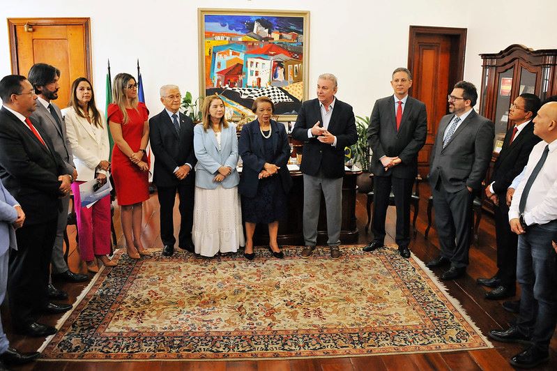  TJMA celebra acordo para solução de conflitos na Comunidade Jussaral - des Froz Sobrinho Ribamar Pinheiro  22/05/2025 Foto horizontal em que o TJMA celebra acordo para solução de conflitos na Comunidade Jussaral. A foto mostra um grupo de pessoas em pé, dispostas em semicírculo, dentro de uma sala. No centro, há um grande tapete estampado no chão de madeira. As pessoas estão vestindo roupas sociais, e há homens e mulheres de diferentes idades. Ao fundo, pode-se ver móveis de madeira escura, como estantes e armários, e um quadro colorido pendurado na parede.