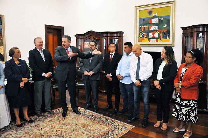 Foto horizontal em que o TJMA celebra acordo para solução de conflitos na Comunidade Jussaral. A foto exibe um grupo de pessoas em pé, dispostas em uma sala de ambiente formal. No centro da imagem, um homem de terno gesticula, explicando algo para os outros. As pessoas ao redor, homens e mulheres de diferentes idades, estão vestindo roupas sociais e algumas parecem estar atentas ao que está sendo dito. O ambiente possui móveis de madeira escura, um tapete estampado no chão e um quadro colorido na parede ao fundo.