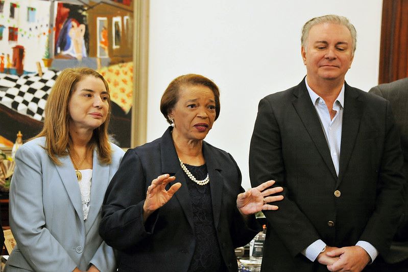 Foto horizontal em que o TJMA celebra acordo para solução de conflitos na Comunidade Jussaral. A foto mostra três pessoas em um ambiente interno. No centro, há uma mulher negra com cabelo curto e escuro, usando um colar de pérolas e um blazer escuro. Ela está gesticulando com as mãos enquanto fala. À esquerda dela, há uma mulher branca com cabelo comprido e castanho, usando um blazer azul-claro e uma blusa branca. Ela está olhando para a frente. À direita da mulher central, há um homem branco com cabelo curto e claro, vestindo uma camisa clara e um blazer escuro. Ele está de pé com as mãos unidas à frente do corpo e os olhos ligeiramente fechados.