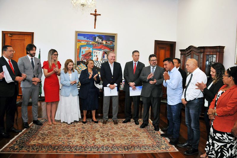 Foto horizontal em que o TJMA celebra acordo para solução de conflitos na Comunidade Jussaral. A foto mostra um grupo de treze pessoas em um ambiente formal, com móveis de madeira escura e quadros na parede. A maioria das pessoas está vestida com trajes sociais. No lado esquerdo da imagem, há um homem usando terno e gravata vermelha, seguido por outros homens e mulheres. Uma mulher no centro do grupo está usando um vestido vermelho vibrante e aplaudindo. Uma outra mulher, mais à direita do centro, está vestindo um blazer azul-claro e uma saia longa branca. No centro da imagem, há uma mulher com cabelo escuro preso, vestindo uma roupa azul-escura e um colar. Ela está aplaudindo. Ao lado dela, há um homem com terno e gravata, segurando papéis. Mais à direita, há um grupo de homens e mulheres. Um homem, de camisa azul-claro e calça jeans, está com as mãos unidas em frente ao rosto, em um gesto que pode indicar agradecimento ou reverência. Uma mulher à direita dele está com as mãos unidas e uma expressão atenta. No fundo da sala, é possível ver um crucifixo na parede, um lustre e uma pintura colorida. O ambiente parece bem iluminado e formal.