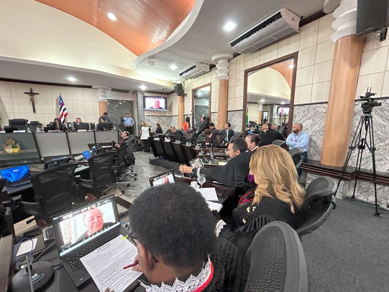 Foto horizontal em que o TJMA define lista tríplice para vaga no TRE destinada a advogadas. A foto mostra o plenário do tribunal, com várias pessoas presentes. Em primeiro plano, de costas, há uma pessoa vestindo uma toga preta com gola branca, olhando para um notebook onde uma videochamada está sendo exibida. Ao lado, outras pessoas também vestindo togas estão sentadas em cadeiras similares. O ambiente é amplo, com mesas de madeira escura dispostas em semicírculo ou em fileiras, e cadeiras estofadas em tons escuros. Há várias pessoas sentadas nas mesas, algumas vestindo togas. Ao fundo, é possível ver um crucifixo na parede, uma bandeira do Maranhão e outra, do Brasil, além de monitores exibindo imagens de pessoas em videochamadas.