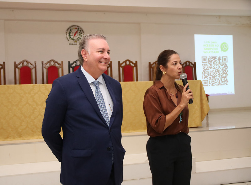 Foto horizontal - TJMA e município entregam 30 registros imobiliários durante curso em Grajaú. A foto mostra duas pessoas em um palco ou área de apresentação. À esquerda, um homem de pele clara veste um terno azul escuro, camisa branca e uma gravata estampada em tons de azul. Ele está sorrindo e olhando para a direita. À direita, uma mulher de pele clara com cabelos castanhos presos veste uma blusa marrom e calças pretas. Ela está segurando um microfone dourado e parece estar falando. Ao fundo, há uma mesa coberta com um tecido estampado em tons de amarelo e branco, com várias cadeiras de madeira escura com detalhes em vermelho atrás dela. À direita, há uma projeção em uma tela branca com um código QR grande, um logotipo do WhatsApp e texto em português.