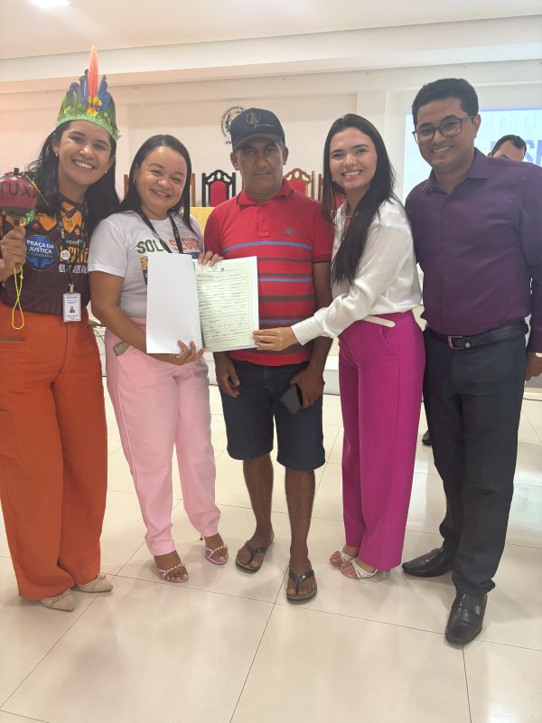 Foto horizontal - TJMA e município entregam 30 registros imobiliários durante curso em Grajaú. Foto mostra cinco pessoas, entre elas o senhor Domingos Ferreira, que recebeu registro do seu imóvel. Três mulheres aparecem na foto com roupas casuais, um delas usando um cocar indígena. Além de seu Domingos, que usa boné, outro homem aparece na foto, feita num auditório.