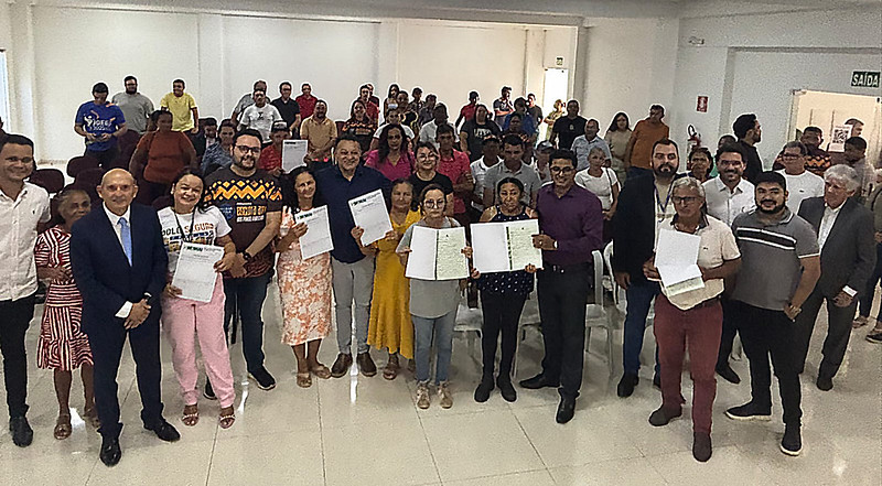 Foto horizontal - TJMA e município entregam 30 registros imobiliários durante curso em Grajaú. A foto mostra um grande grupo de pessoas reunidas em um espaço amplo e bem iluminado, posando para uma foto. Há aproximadamente 30 a 40 pessoas, incluindo homens e mulheres de diversas idades e aparências. Na frente, várias pessoas seguram o que parecem ser certificados ou documentos, alguns com sorrisos no rosto. Um homem careca de pele clara, vestindo um terno azul e gravata azul, está posicionado ligeiramente à frente e ao centro do grupo. As pessoas na foto vestem uma variedade de roupas casuais, incluindo camisetas, camisas, vestidos e calças. A composição do grupo sugere uma diversidade de idades e possivelmente origens étnicas. O ambiente parece ser um salão de eventos ou um espaço comunitário com paredes brancas e iluminação artificial. Ao fundo, há uma porta com a palavra "SAÍDA" escrita em cima, indicando uma saída de emergência. Há também outras portas e o que parece ser um bebedouro visível ao fundo.
