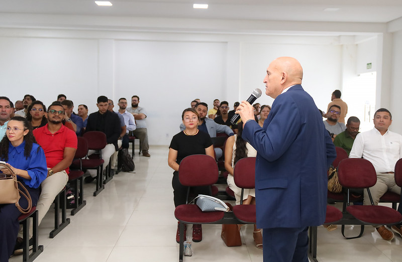 Foto horizontal - TJMA e município entregam 30 registros imobiliários durante curso em Grajaú. A foto mostra um homem calvo de pele clara, vestido com um terno azul e uma camisa branca, falando em um microfone para uma plateia sentada em cadeiras vermelhas. O homem está de costas para a câmera, voltado para a plateia. A plateia é composta por diversas pessoas, homens e mulheres, de diferentes aparências e idades. Eles estão sentados e olhando na direção do orador. As roupas da plateia variam de casuais a um pouco mais formais. O ambiente é um auditório pequeno, com paredes claras e iluminação artificial vinda do teto. Há janelas com cortinas claras ao fundo.