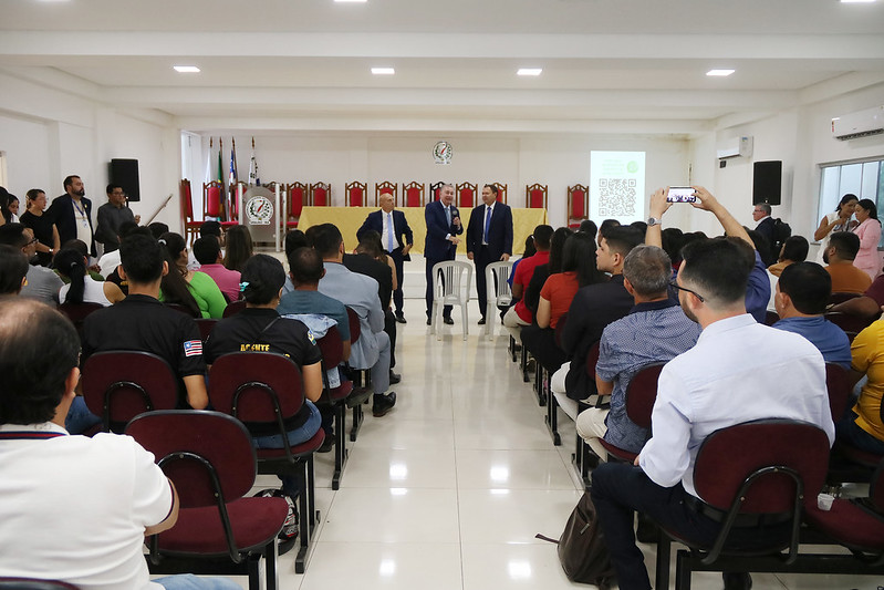 Foto horizontal - TJMA e município entregam 30 registros imobiliários durante curso em Grajaú. A foto mostra um auditório com várias pessoas reunidas. Há um palco elevado ao fundo, onde quatro homens estão em pé. Três dos homens vestem ternos escuros e camisas claras, enquanto o quarto homem, posicionado mais à esquerda no palco, veste uma camisa clara e calças escuras. Eles parecem estar falando ou apresentando algo, possivelmente em um evento formal. Na plateia, várias pessoas estão sentadas em cadeiras escuras, voltadas para o palco. As pessoas na plateia vestem roupas casuais e parecem estar prestando atenção ao que está acontecendo no palco. Algumas pessoas estão olhando para o palco, enquanto outras parecem estar interagindo ou olhando para seus dispositivos. Ao fundo, no palco, há uma mesa com cadeiras vermelhas atrás, e bandeiras do Brasil e do Maranhão são visíveis à esquerda. Um símbolo ou brasão oficial também está presente no centro do palco. À direita do palco, há uma projeção em uma tela branca com um código QR e outras informações. Caixas de som pretas estão montadas nas paredes laterais.