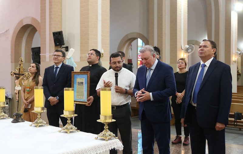 Foto horizontal. TJMA entrega registro imobiliário de Catedral de Grajaú. A foto mostra um grupo de sete pessoas dentro de uma igreja, reunidas em frente a uma mesa coberta com uma toalha branca, onde há grandes velas acesas em castiçais dourados. Da esquerda para a direita: Uma mulher de pele clara com cabelos longos e escuros veste um vestido claro com detalhes em laranja e está olhando para a direita. Um homem de pele clara veste um terno azul escuro e gravata azul, olhando para a frente. Um homem de pele clara veste uma batina preta e óculos, segurando um objeto emoldurado e olhando para a frente. Um homem de pele clara veste uma camisa branca de manga curta e óculos, falando em um microfone preto e olhando para baixo. Um homem de pele clara veste um terno azul e gravata estampada, com as mãos juntas à frente e olhando para baixo. Uma mulher de pele clara com cabelos escuros presos veste uma blusa preta e calças escuras, olhando para a direita. Um homem de pele clara veste um terno azul e gravata azul, olhando para a esquerda. Ao fundo, a igreja apresenta arcos arquitetônicos, pilares e iluminação suave. Uma grande cruz dourada é visível à esquerda. Há imagens religiosas em nichos nas paredes.
