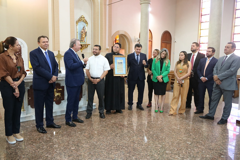 TJMA entrega registro imobiliário de Catedral de Grajaú. A foto mostra um grupo de aproximadamente 11 pessoas posando para uma foto dentro de uma igreja. À esquerda, uma mulher com cabelos castanhos e uma blusa marrom está olhando para a direita. Ao seu lado, um homem de terno azul e gravata azul está olhando para a frente com um leve sorriso. Um homem de terno azul e gravata estampada está ao seu lado, falando em um microfone preto. No centro, um homem com uma camisa branca de manga curta e óculos está sorrindo. Ao seu lado, um homem vestido com roupas clericais pretas está segurando um objeto emoldurado e sorrindo. Um homem de terno azul escuro está ao lado dele, olhando para o objeto emoldurado. À direita, uma mulher com uma blusa verde e saia preta está sorrindo. Ao seu lado, uma mulher com um vestido claro está sorrindo. Um homem de terno escuro e gravata clara está ao lado dela, olhando para a frente. Outros homens de terno completam o grupo à direita. O altar da igreja é visível ao fundo, com nichos contendo estátuas religiosas. A arquitetura da igreja apresenta arcos e janelas com vitrais. O chão é de ladrilho em tons de marrom e bege.