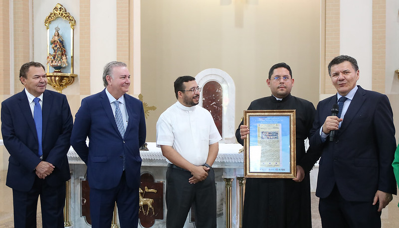 Foto horizontal. TJMA entrega registro imobiliário de Catedral de Grajaú. A foto mostra cinco homens em pé dentro de uma igreja, posando para uma foto em frente ao altar. Da esquerda para a direita: Um homem de pele clara veste um terno azul escuro e uma gravata azul. Ele está olhando para a frente com um leve sorriso. Um homem de pele clara veste um terno azul com uma gravata estampada em tons de azul. Ele está olhando para a frente com um sorriso. Um homem de pele clara veste uma camisa branca de manga curta e óculos. Ele está sorrindo e olhando para a frente com as mãos juntas à frente do corpo. Um homem de pele clara veste uma batina preta e óculos. Ele está segurando um objeto emoldurado, possivelmente um certificado ou uma placa, e sorrindo para a câmera. Um homem de pele clara veste um terno azul escuro e está falando em um microfone preto. Ele está olhando para a direita. O altar atrás deles é branco com detalhes dourados. Há nichos nas paredes da igreja com estátuas religiosas. A arquitetura da igreja inclui arcos e pilares em tons claros.