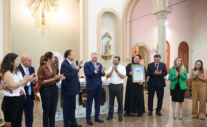 TJMA entrega registro imobiliário de Catedral de Grajaú. A foto mostra um grupo de aproximadamente 12 pessoas dentro de uma igreja, em frente ao altar, aplaudindo. À esquerda, uma mulher com cabelos escuros e uma blusa estampada está aplaudindo. Ao seu lado, um homem calvo com um terno azul e uma gravata azul também está aplaudindo. Uma mulher com uma blusa marrom e calças pretas está ao lado dele, também aplaudindo. Um homem com um terno azul e uma gravata azul está no centro esquerdo, aplaudindo. Ao seu lado, um homem com um terno azul está sorrindo e aplaudindo. No centro, um homem com uma camisa branca de manga curta e óculos está sorrindo e aplaudindo. Ao seu lado, um homem vestido com roupas clericais pretas está segurando um objeto emoldurado e sorrindo enquanto aplaude. À direita, um homem com um terno azul escuro e óculos está aplaudindo. Uma mulher com um blazer verde e saia preta está ao seu lado, aplaudindo. Outras pessoas, incluindo mulheres com roupas claras, também estão aplaudindo. O altar atrás deles é branco com detalhes dourados. A arquitetura da igreja inclui arcos e pilares em tons claros. Uma grande cruz dourada é visível ao fundo.