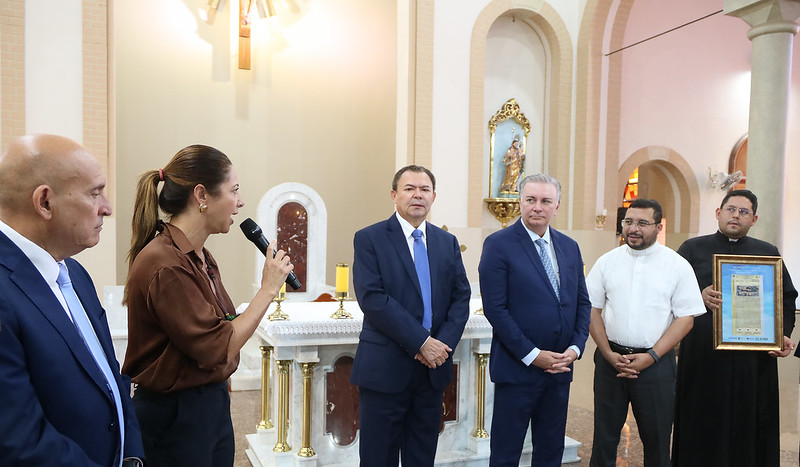 Foto horizontal. TJMA entrega registro imobiliário de Catedral de Grajaú - diretora do TJMA. A foto mostra um grupo de seis pessoas dentro de uma igreja, reunidas em frente ao altar. À esquerda, um homem calvo de pele clara veste um terno azul e olha para a mulher ao seu lado. A mulher ao seu lado, com cabelos castanhos presos, veste uma blusa marrom e está falando em um microfone preto. No centro, dois homens de pele clara vestem ternos azuis e camisas brancas. Eles estão lado a lado, olhando para a frente. À direita, dois homens vestidos com roupas clericais estão presentes. O homem mais próximo veste uma camisa branca de manga curta e óculos. O homem atrás dele veste uma batina preta e também usa óculos. Ele está segurando um objeto emoldurado com documento.