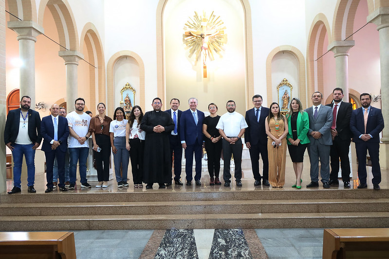TJMA entrega registro imobiliário de Catedral de Grajaú. A foto mostra um grupo de aproximadamente 19 pessoas posando para uma foto dentro de uma igreja. O grupo é composto por homens e mulheres de diversas idades. No centro, um homem vestido com roupas clericais pretas se destaca. À sua esquerda e direita, há outros homens e mulheres, alguns vestindo roupas formais como ternos e blazers, enquanto outros vestem roupas mais casuais, incluindo camisetas com estampas. O interior da igreja apresenta arcos arquitetônicos, pilares e iluminação suave. Ao fundo, acima do grupo, há uma grande cruz dourada com a figura de Cristo. Há também nichos nas paredes com imagens religiosas. Os degraus de mármore onde o grupo está posando levam a uma área mais elevada da igreja. Bancos de madeira escura são visíveis em primeiro plano, fora do foco principal.